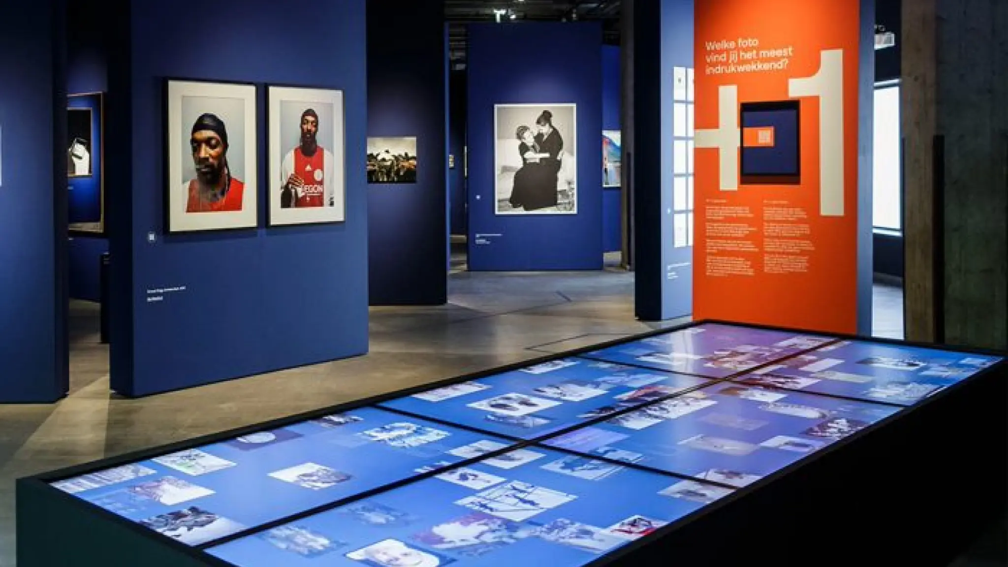 Eregalerij van het Nederlands Fotomuseum. Op blauwe wanden hangen grote foto's. Op de voorgrand zijn led-schermen te zien waarop bezoekers foto's kunnen aanklikken. Op een oranje paneel staat de vraag 'Welke foto vind jij het meest indrukwekkend?'