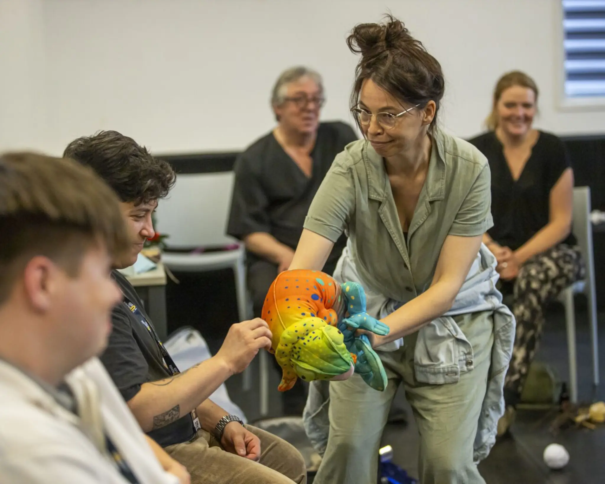 Een workshopleider van Concrete Youth houdt een handpop van een kameleon vast. Een van de deelnemers steekt zijn hand uit om aan de kameleon te voelen.