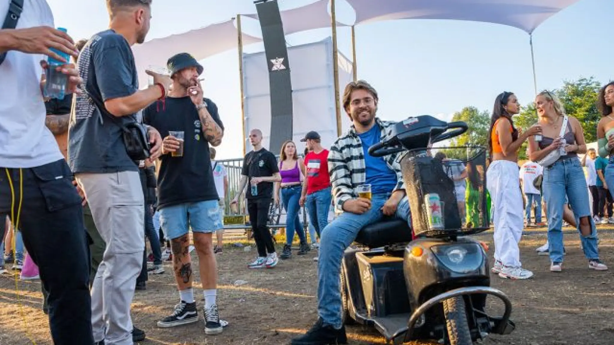 Een jonge man zit op een scootmobiel op een festivalterrein. Hij en andere festivalgangers om hem heen hebben drankjes in hun hand en genieten van de sfeer.