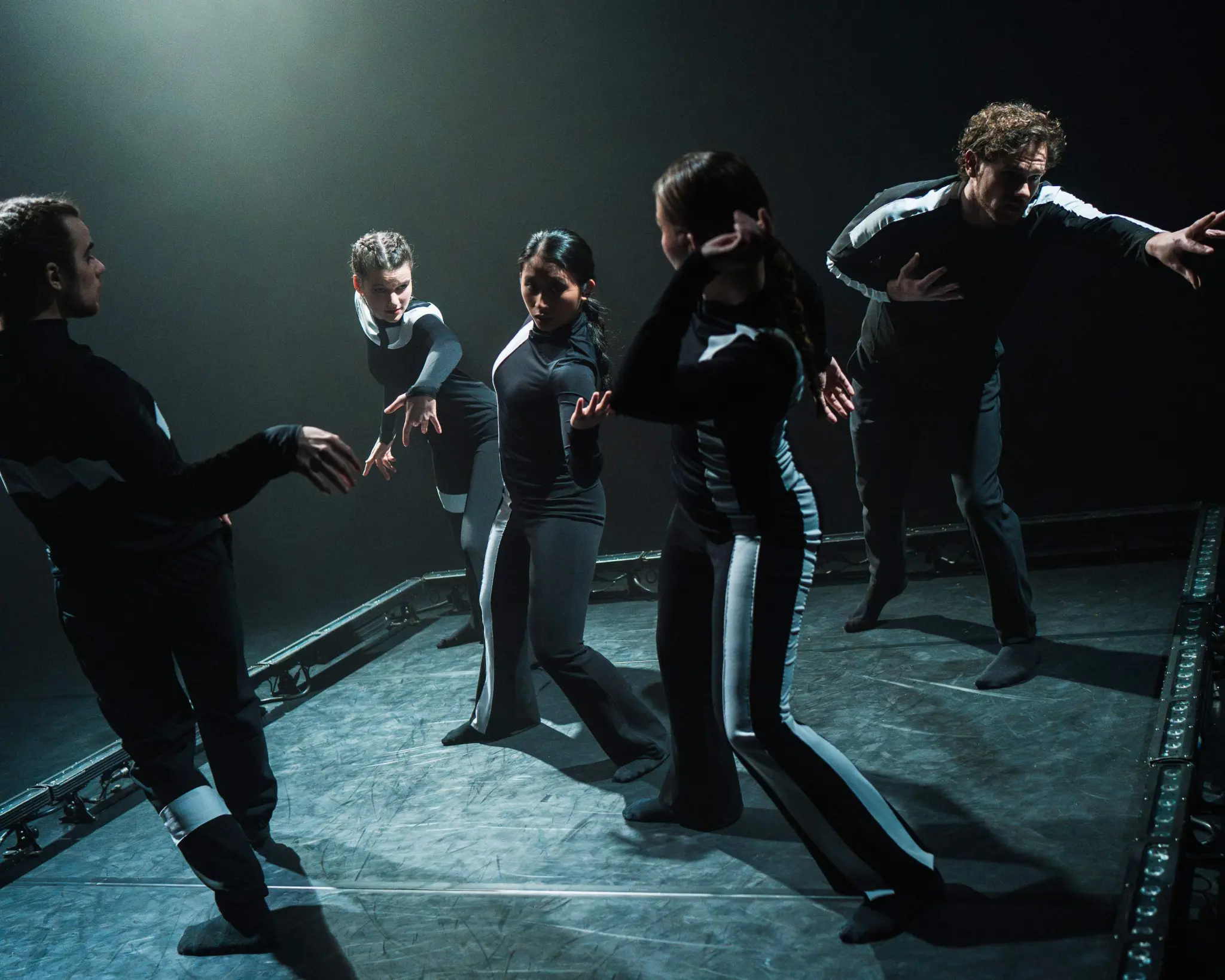 Vijf dansers bewegen op een donker podium, slechts verlicht door een spotlight. Ze dragen zwarte pakken met reflecterende zilveren strepen.
