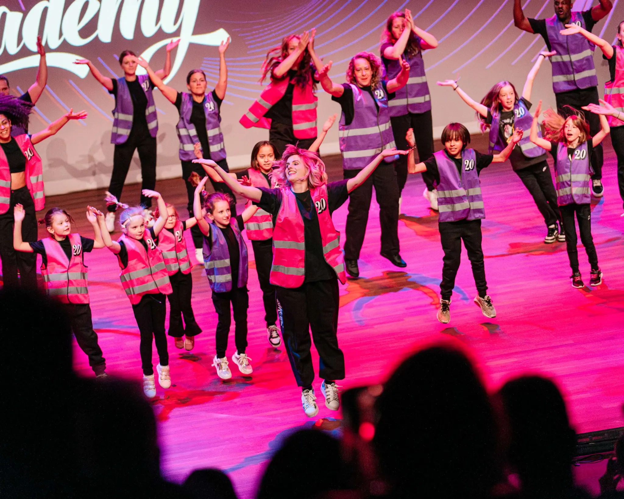 Een groep kinderen geeft een dansvoorstelling. Ze dragen roze en paarse hesjes met het nummer 20 erop. Ze springen met hun handen in de lucht.