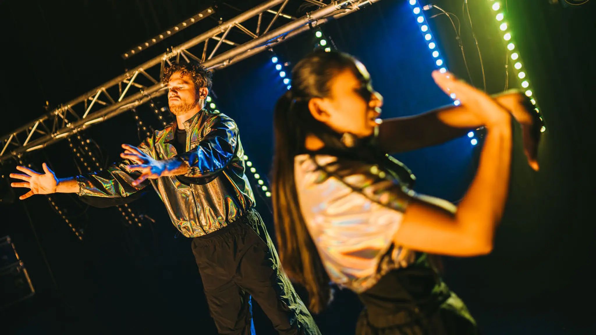 Een man en een vrouw dansen op een podium, in een gouden licht. Ze dragen een zwarte broek en goudkleurig jasje. Ze maken allebei gebaren met hun handen.