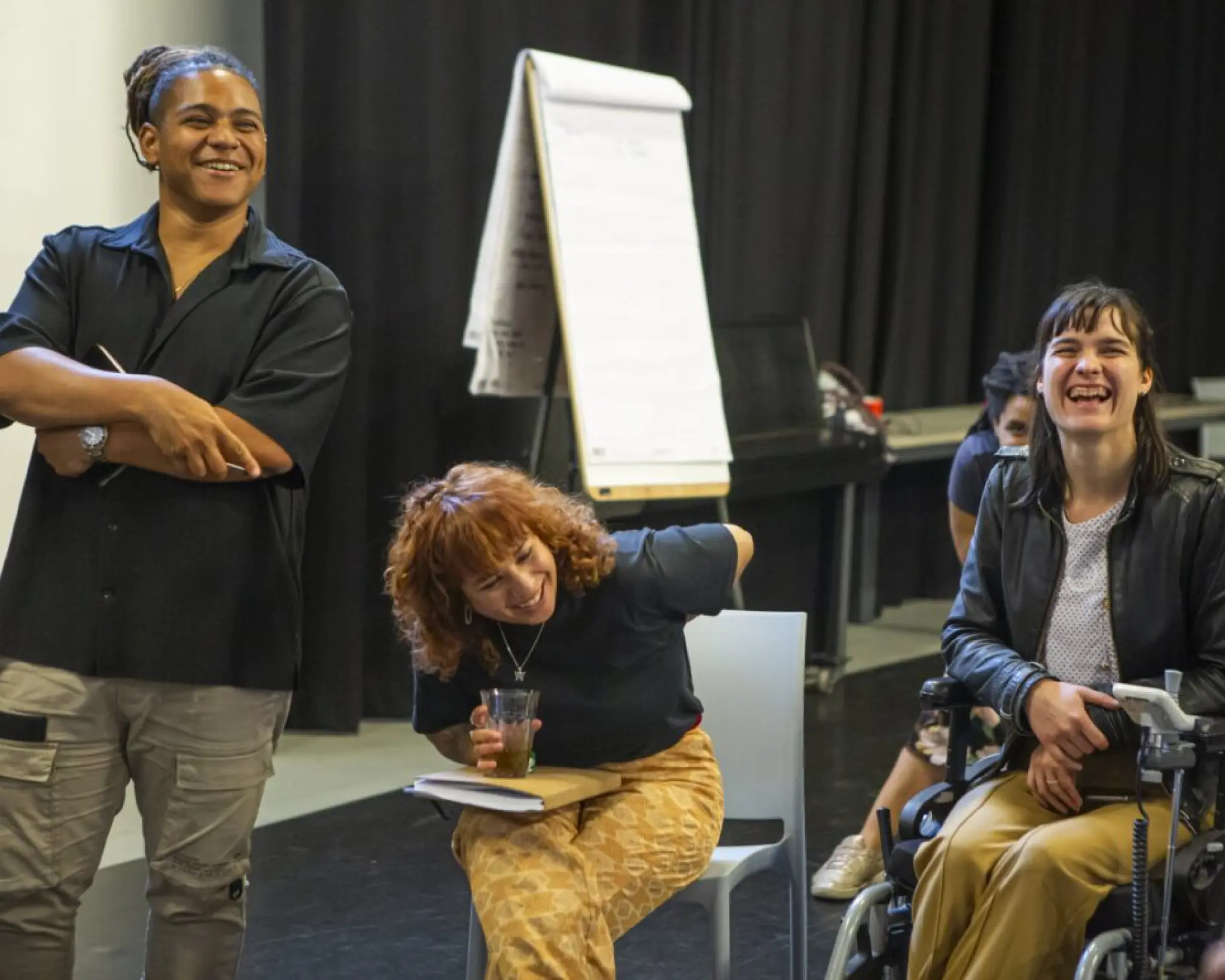 Drie deelnemers van een workshop van Concrete Youth naast elkaar. Links staat een jonge man, in het midden zit een honge vrouw op een stoel.  Rechts zit een jonge vrouw in een rolstoel. Alle drie lachen ze. Op de achtergrond staat een flip-over.
