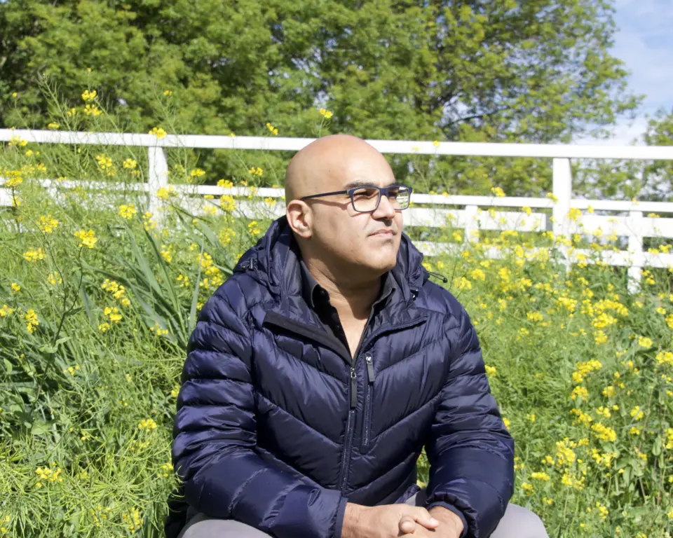 Mohamed Bouzia zit gehurkt in een weiland met gele bloemen. Achter hem is een wit hekje en een boom te zien.