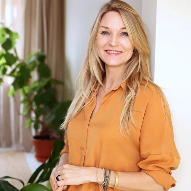 Eva Kuhlemeier, leunend tegen een witte muur in een huiskamer. Op de achtergrond gezellige planten
