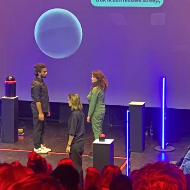 Publiek zit op rode stoelen in een halfronde zaal rondom een theatervloer. Op de vloer staan vier personen in een vierkant, waarbij steeds 2 personen elkaar aankijken.