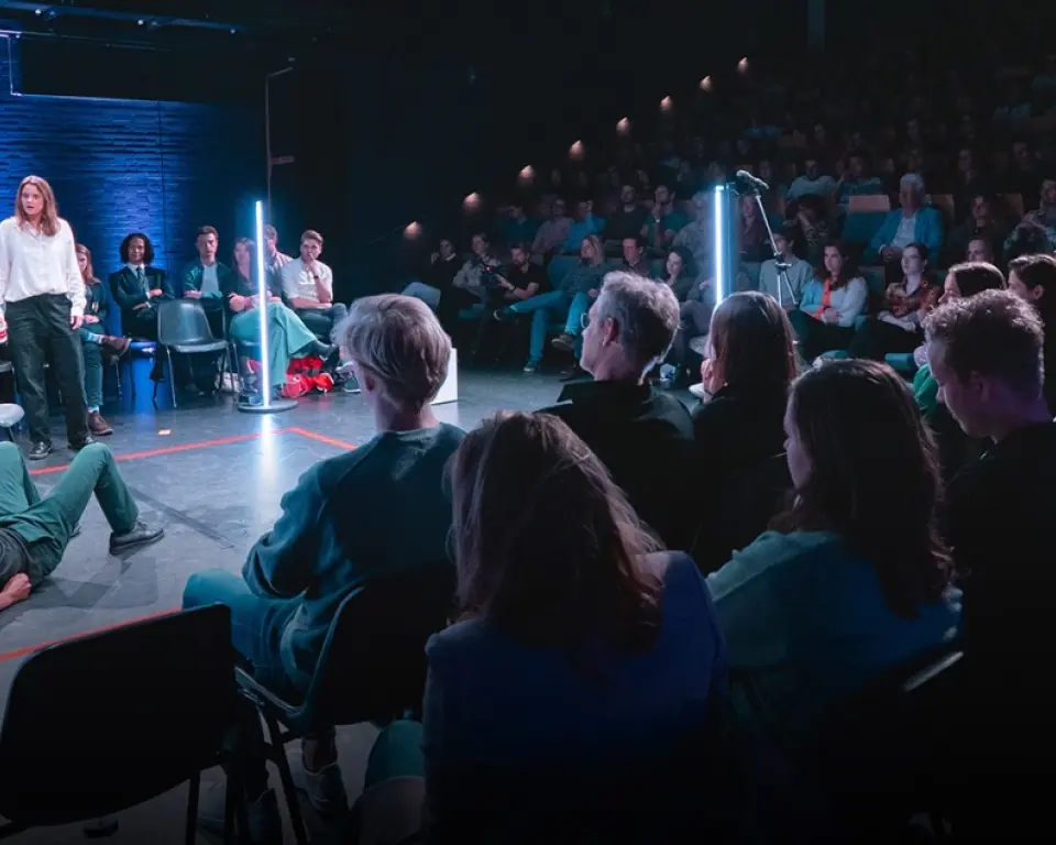 In een donkere zaal zit het publiek op stoelen rondom een vloer. Er zijn drie mensen op de vloer: 1 staat, 1 zit op een stoel en 1 ligt achteroverleunend op de grond.