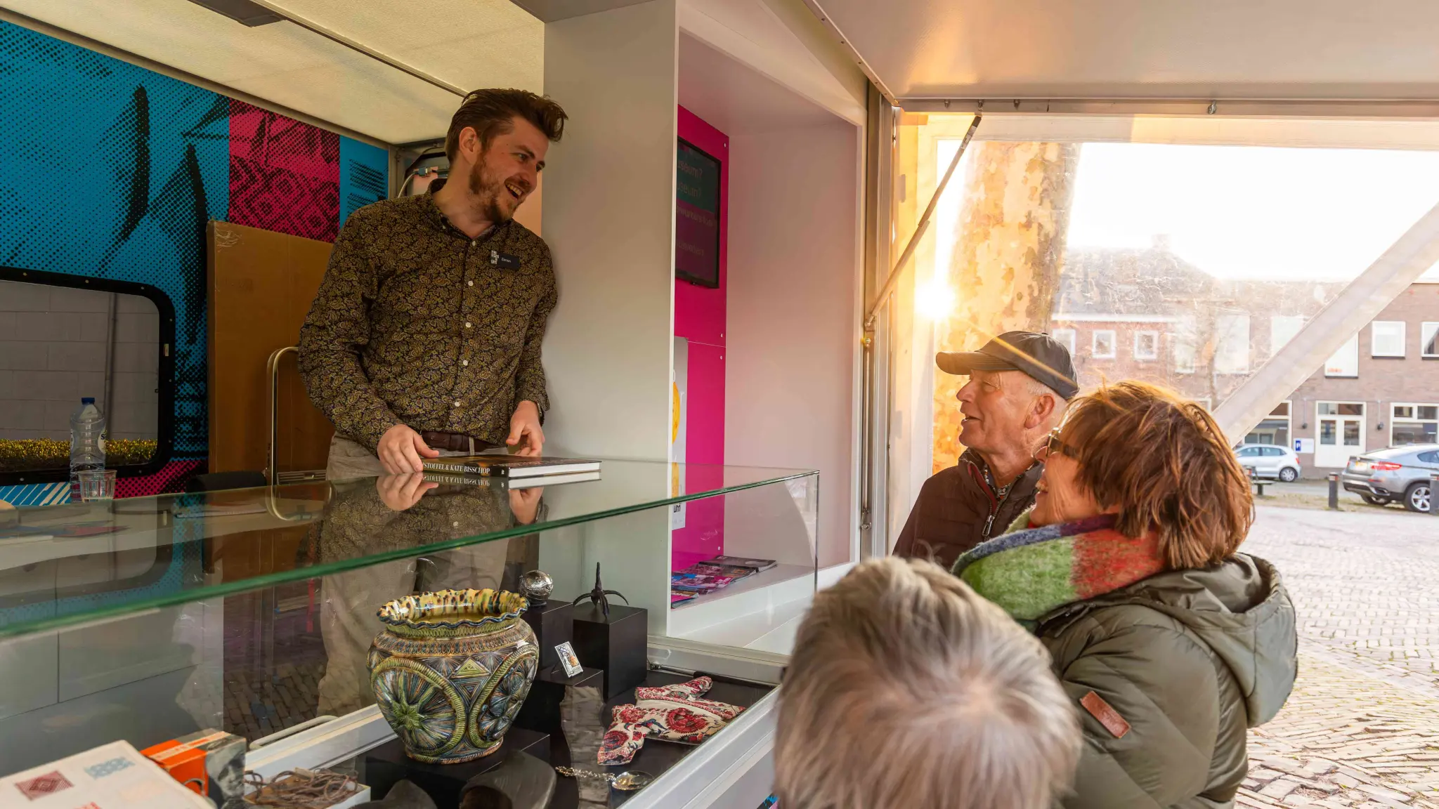 Een medewerker van het museum staat in de viskraam en praat met bezoekers over de kunsttukken in de vitrine. Er liggen onder meer een grote aardewerken pot, een zilveren lepel en een paar oude schoenen.