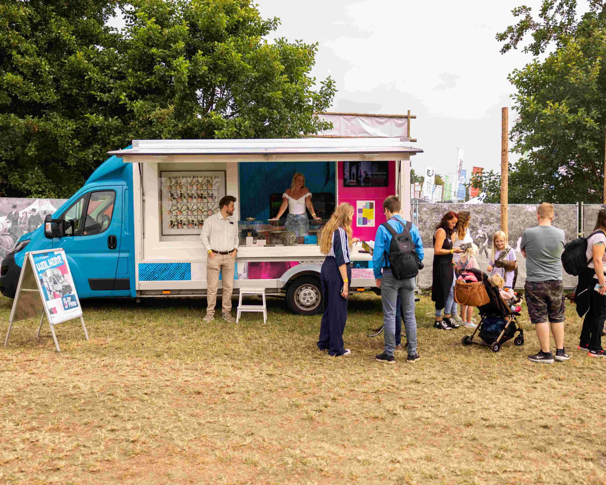 De viskraam met kunst op een grasveld van een festivalterrein. Bezoekers bladeren door de catalogus en foldertjes. Twee medewerkers van het museum staan in en naast de bus.