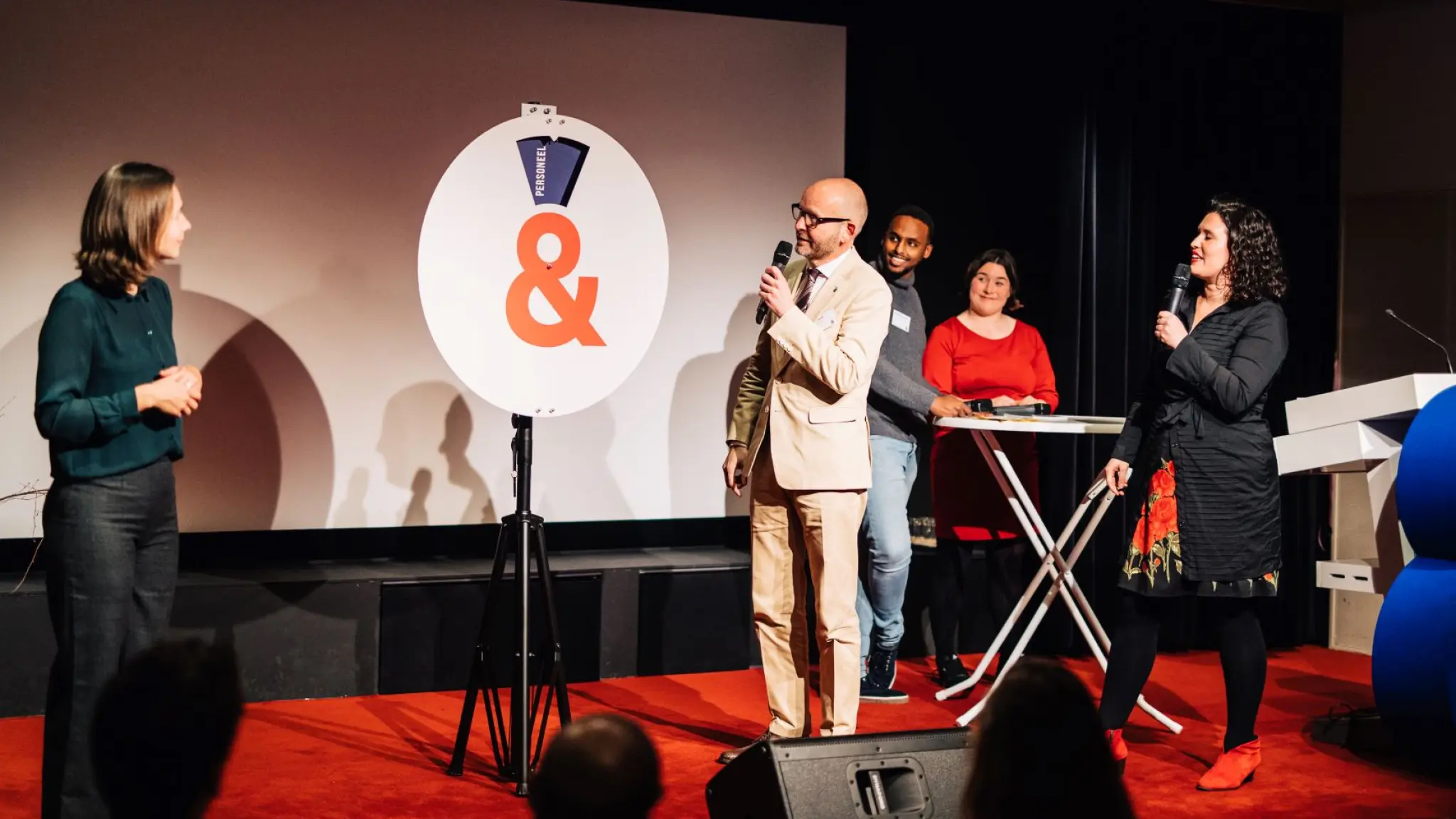 Vijf mensen met microfoons in hun handen staan rondom een groot pim-pam-pet bord op het podium. De uitkomst toont de categorie personeel.