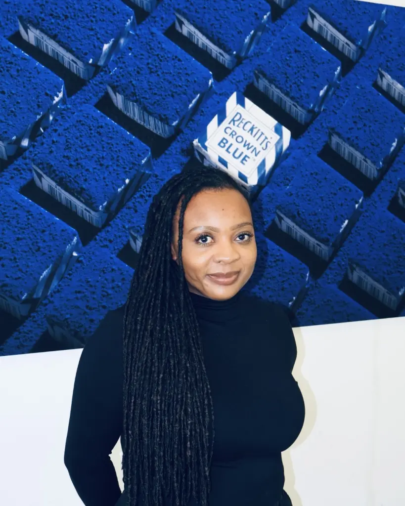 Cheryl Mak heeft haar haar in lange dreadlocks of vlechtjes en ze draagt een zwarte coltrui. Ze staat voor een grote afbeelding van blauwe blokken met op een ervan de tekst Reckitt's Crown Blue. Ze kijkt recht in de camera en glimlacht licht.