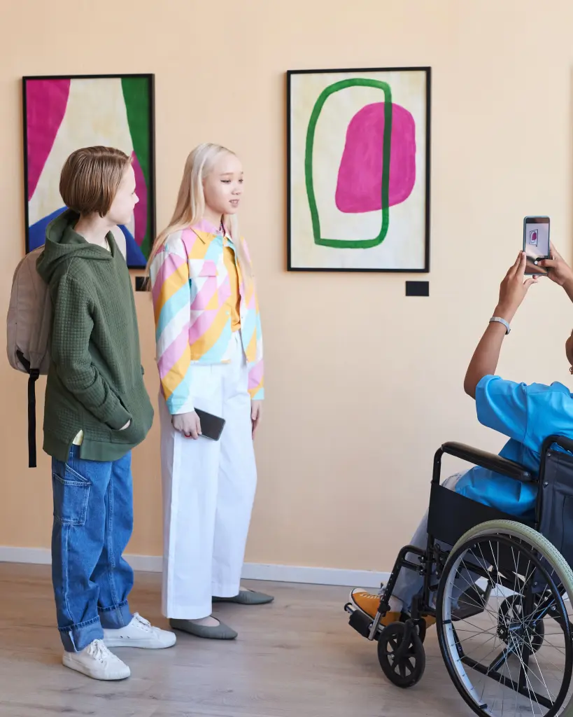 Drie tieners die naar een kunstwerk kijken. Eén tiener zit in een rolstoel met haar rug naar de camera toe. De tiener maakt een foto van het kunstwerk.