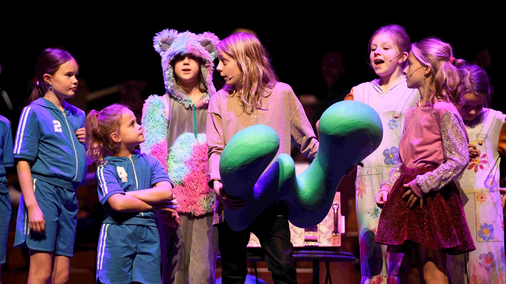 Kinderen geven een voorstelling. Een meisje in het midden draagt een groene letter W. Twee meisjes dragen blauwe sportkleding. Twee meisjes dragen overalls met bloemen. Een meisje heeft een berenpak aan en een ander meisje draagt een roze glittershirt en glitterrok.
