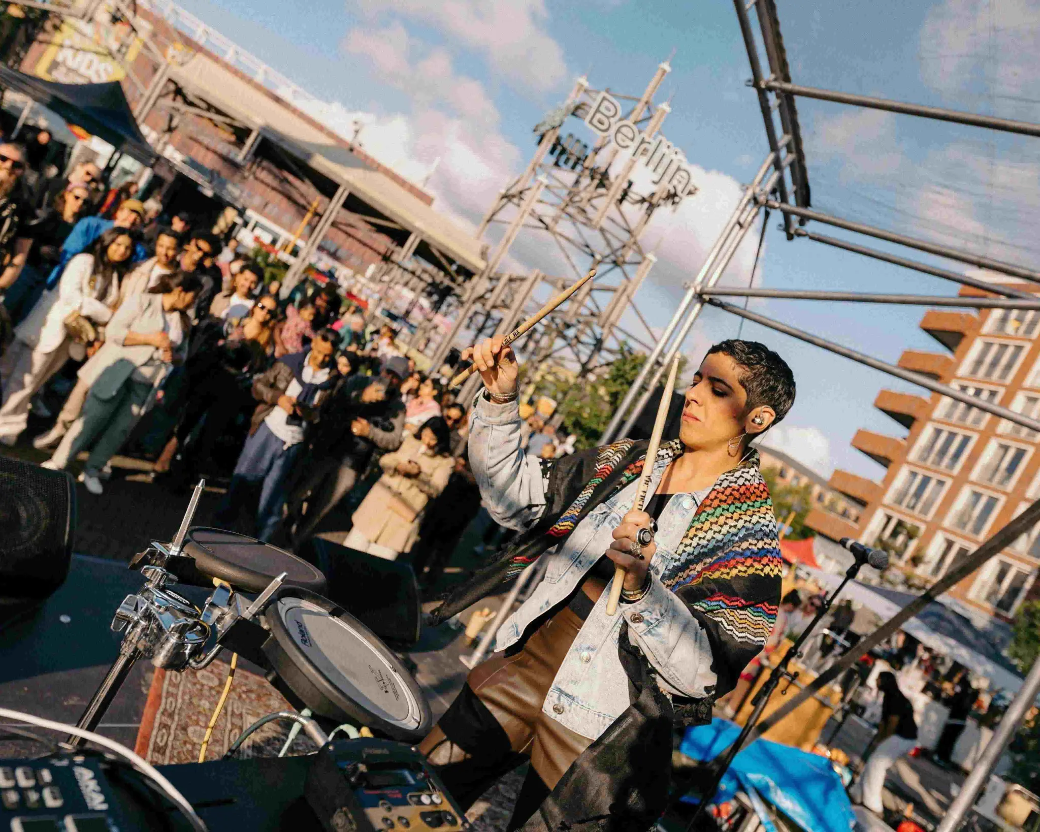 Een vrouw met kort haar een een traditionele sjaal om staat als achter een drumstel op het podium. Ze heeft haar handen omhoog met twee drumsticks. Op de achtergrond staat publiek te luisteren.