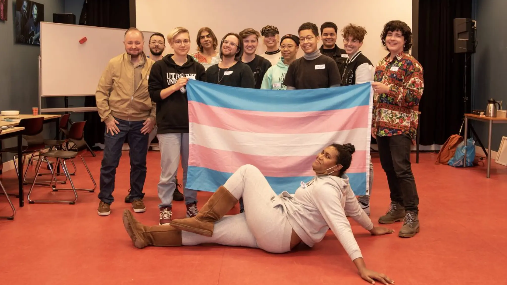 Groep mensen poseert in een ontmoetingsruimte achter een grote transgendervlag. Op de voorgrond ligt één deelnemer voor de vlag op de vloer. Op de achtergrond zijn tafels, stoelen en een presentatiescherm zichtbaar.