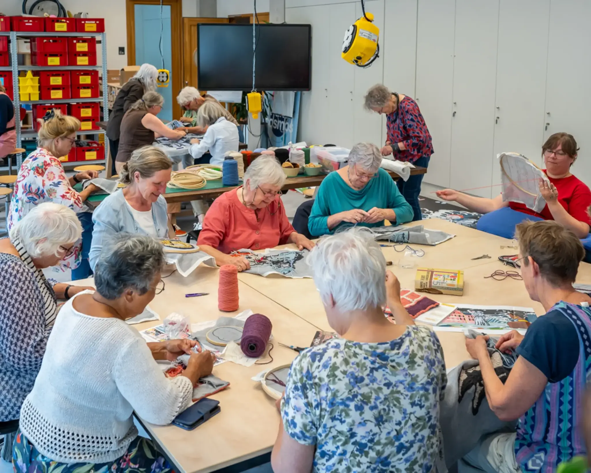 Een groep mensen is aan het borduren in een werkruimte. Ze zijn verdeeld over verschillende tafels. Op de achtergrond een stellingkast met kratten vol materialen.