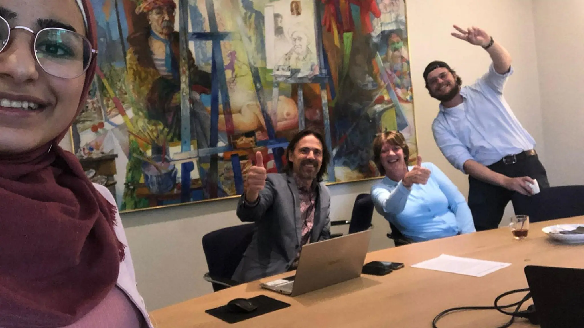 Vier mensen poseren lachend in een vergaderruimte. Op de voorgrond maakt een persoon een selfie, terwijl drie anderen aan een vergadertafel zitten of staan en een duim omhoog of vredesteken geven. Op tafel staan een laptop, papieren en koffiekopjes. Op de achtergrond hangt een groot kleurrijk schilderij.