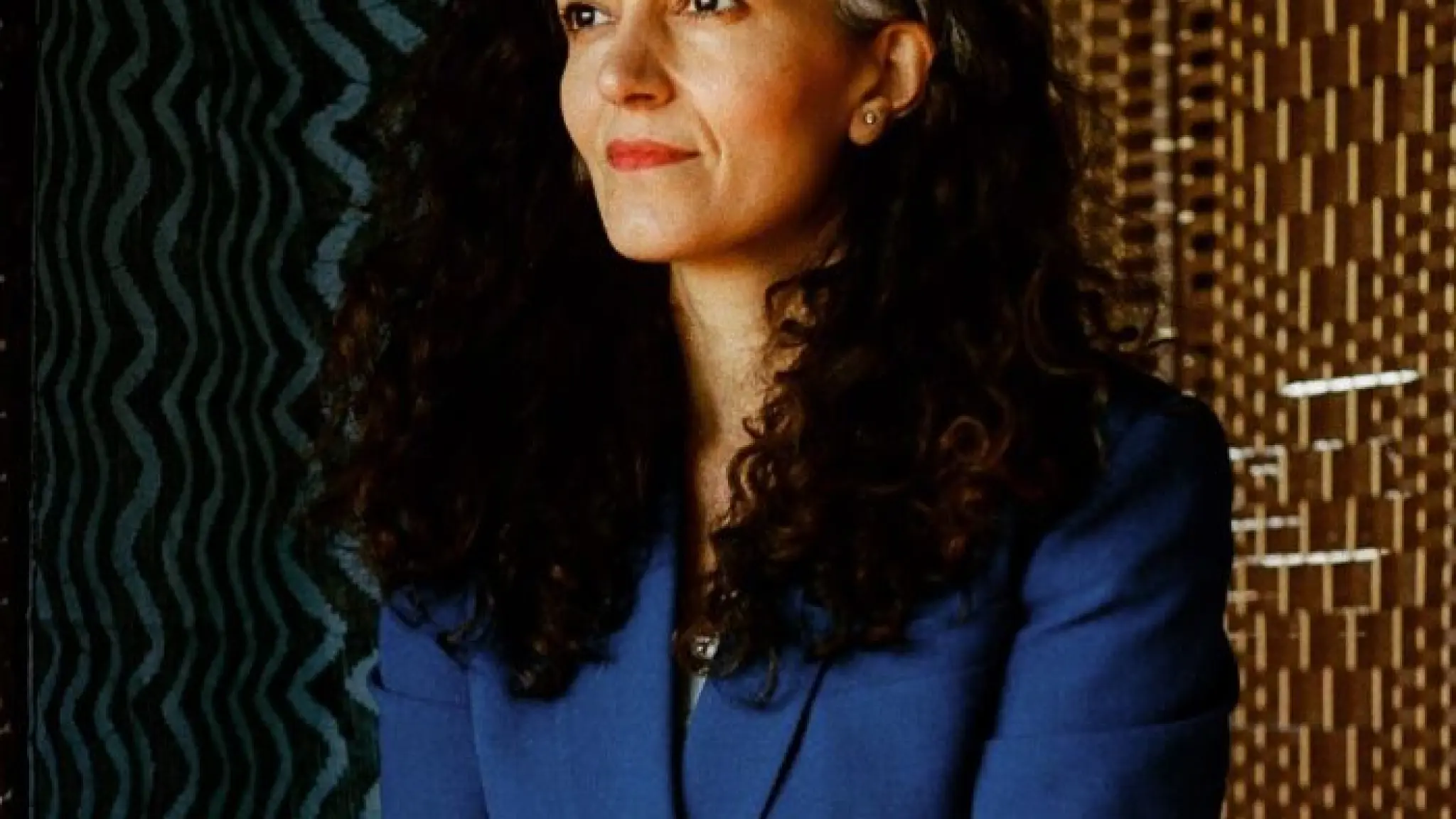 Gonca Yalçıner staatvoor een kamerscherm en een blauw gegolfd doek. Ze draagt een blauwe blazer en heeft haar lange zwarte krullen los hangen.
