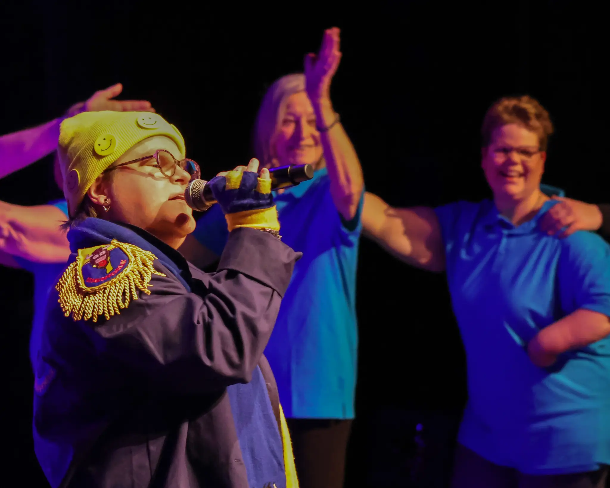 Een vrouw met een gele muts met smilies zicht in een micrfoon. Op de achtergornd doen mensen met een blauwe polo aan de polonaise.