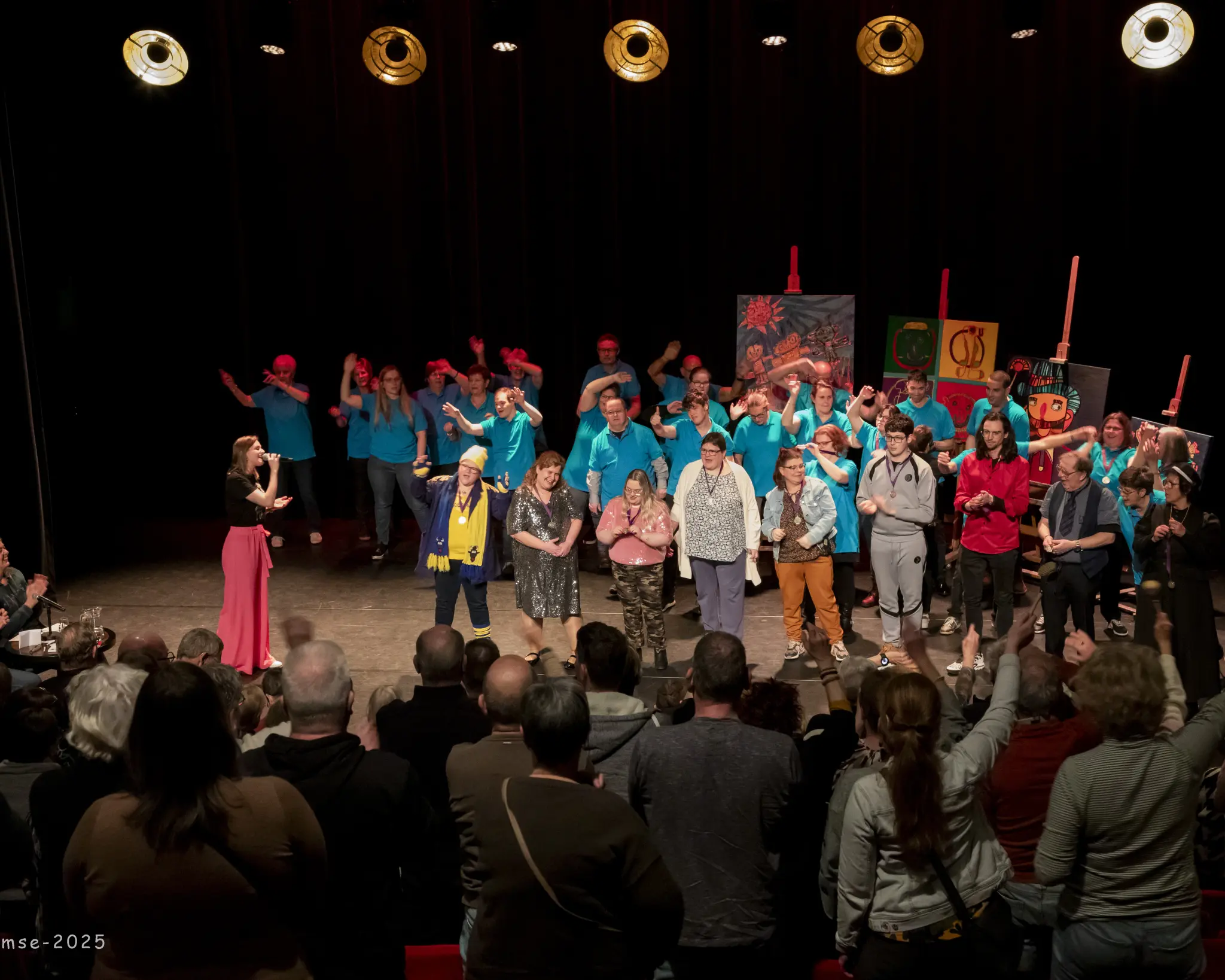 Een groep deelnemers aan een voorstelling staat allemaal bijeen op het podium. Ze worden toegezongen door een vrouw aan de linkerkant. Deelnemers en publiek bewegen met hun handen in de lucht mee op de muziek.