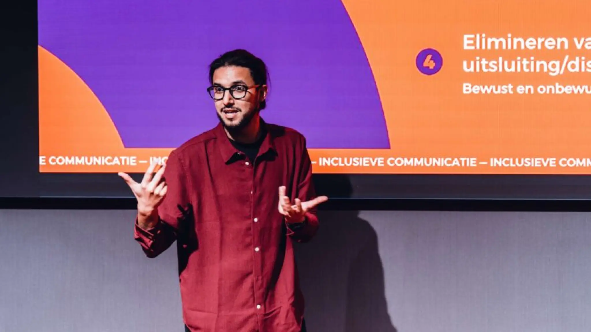 Hamza Ouamari staat voor een presentatiescherm. Hierop zijn paarse en oranje vormen te zien en de tekst Inclusieve communicatie.