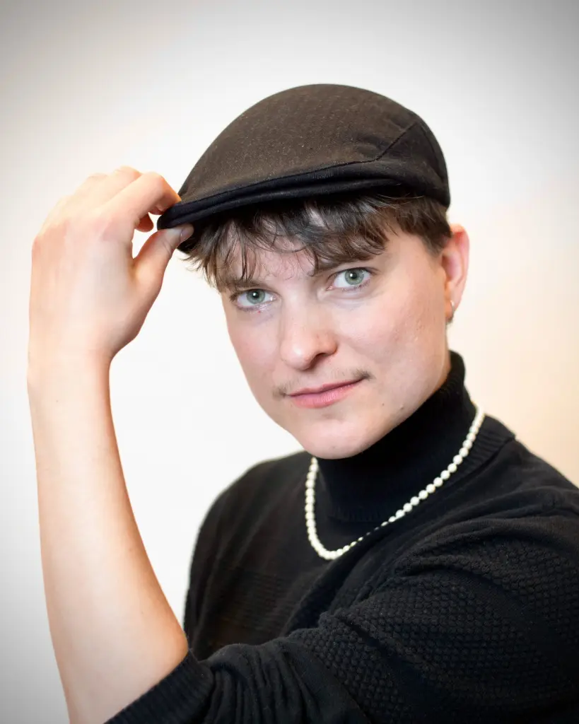 Kir draagt een zwarte coltrui, een parelketting en een baret. Kir kijkt vriendelijk naar de camera terwijl hen met diens hand de voorste rand van de baret vastheeft.