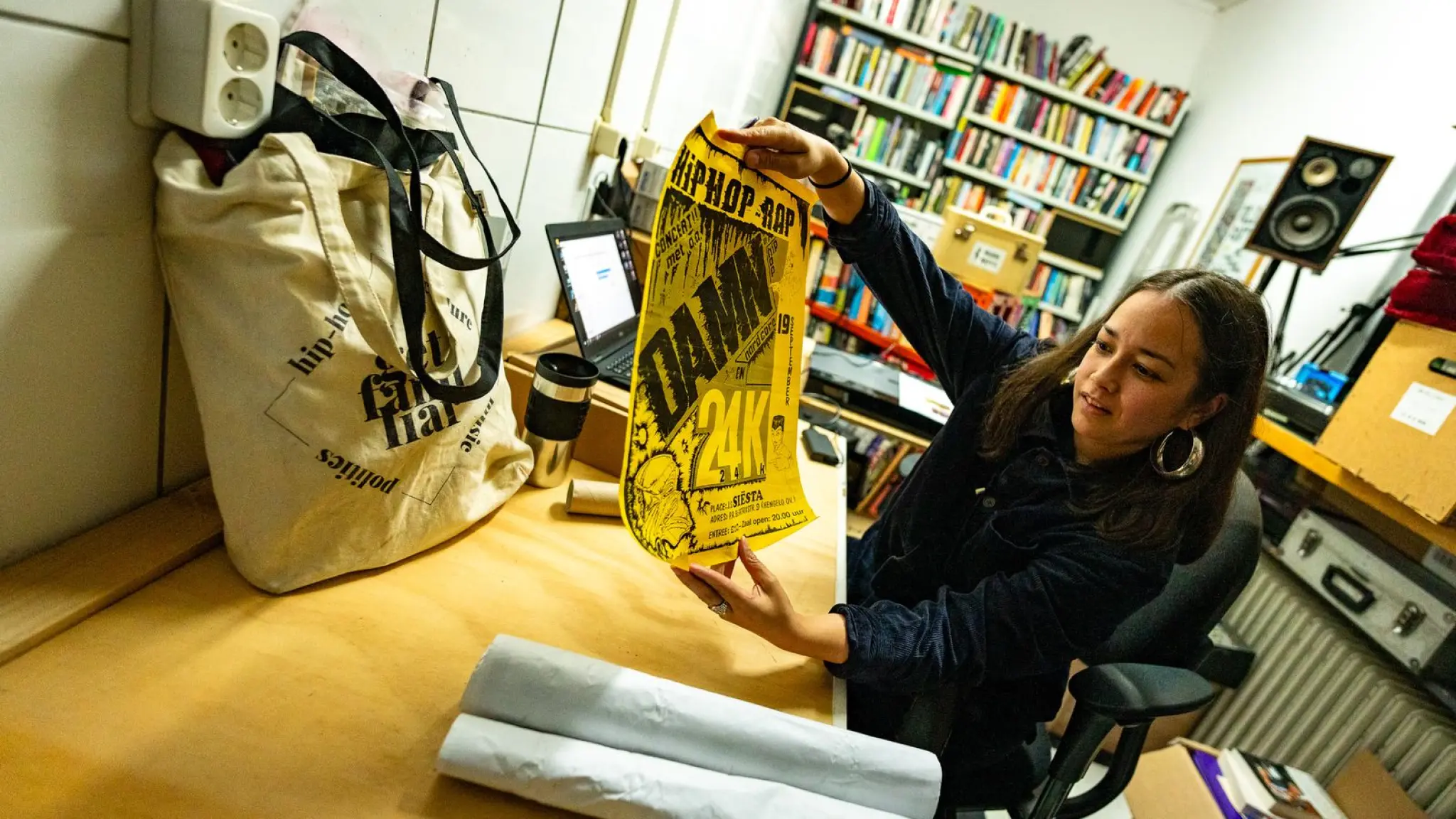 Een vrouw houdt een zwart-wit poster omhoog, gedrukt op geel papier. Bovenaan staat Hiphop-Rap. Eronder staat DAMN 24K.