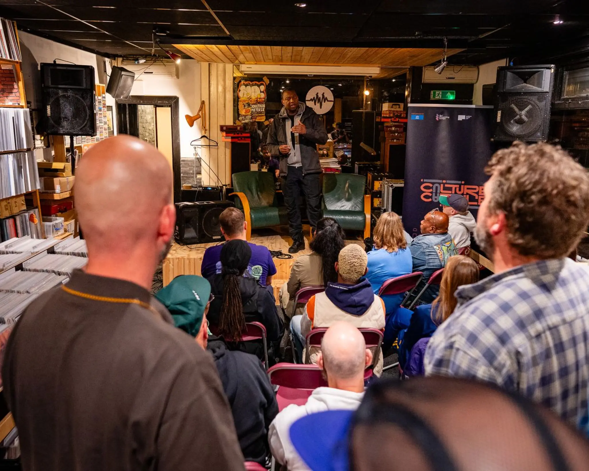 Sherlock Telgt staat op het podium en houdt een microfoon vast. Het publiek zit op stoelen en staat er omheen. Links zijn vele bakken met LP's te zien.
