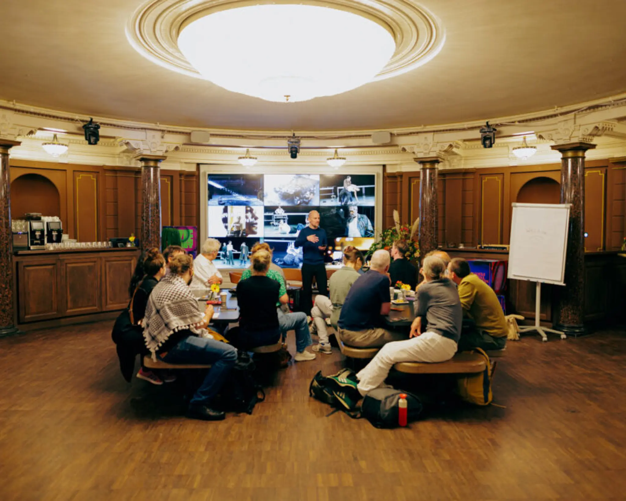 Deelnemers aan een break-out sessie zitten aan tafeltjes in een ronde foyer met marmeren zuilen. De sessieleider staat voor presentatiescherm. Rechts staat een flip-board.