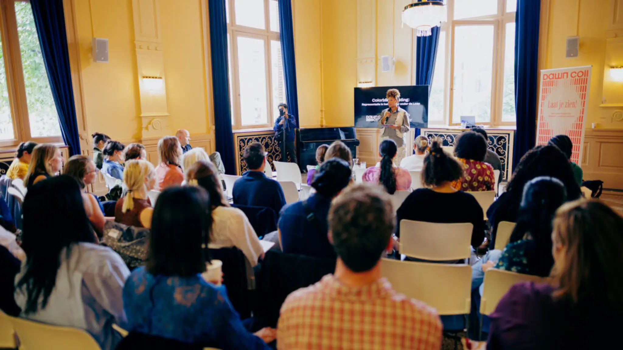 In een gele zaal met blauwe gordijnen luisteren bezoekers aan de sessie van Dawn Collective aandachtig naar de spreker. De spreker staat met een microfoon in haar hand vooraan de zaal. De bezoekers zijn van achteren gefotografeerd.