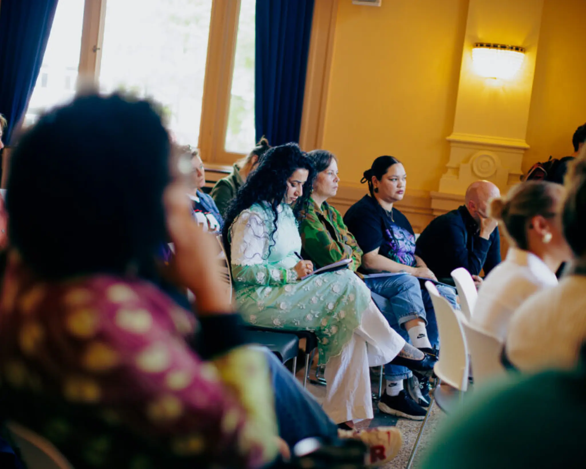 Bezoekers aan de sessie van Dawn Collective luisteren aandachtig naar een spreker buiten beeld. Een vrouw in het midden van de foto maakt aantekeningen op een notitieblok op schoot.