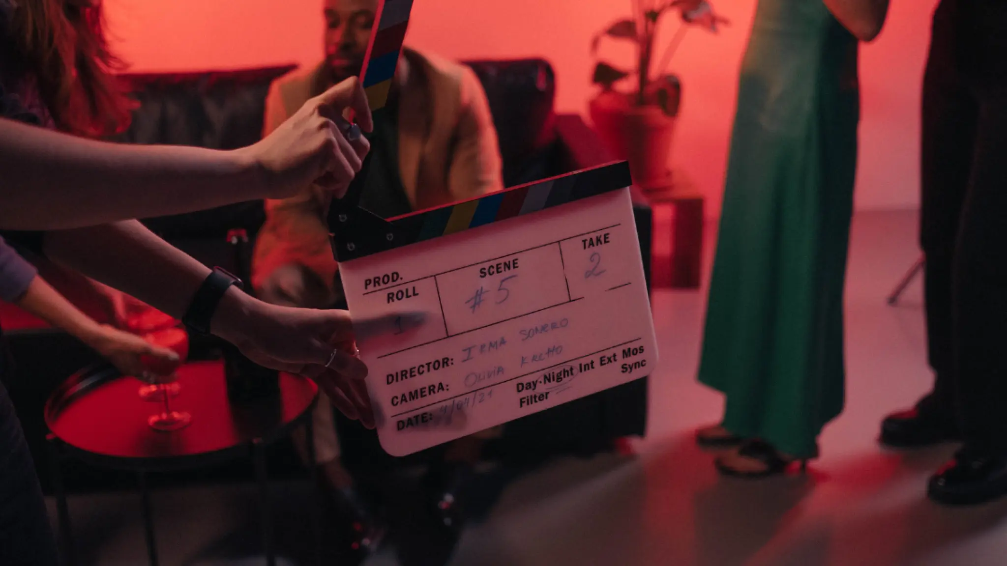 Een paar handen houden een filmklapper in beeld waarop aangegeven staat dat Scene #5, take 2 opgenomen gaat worden. Op de achtergrond zijn staande en zittende acteurs te zien in een rood warm licht.