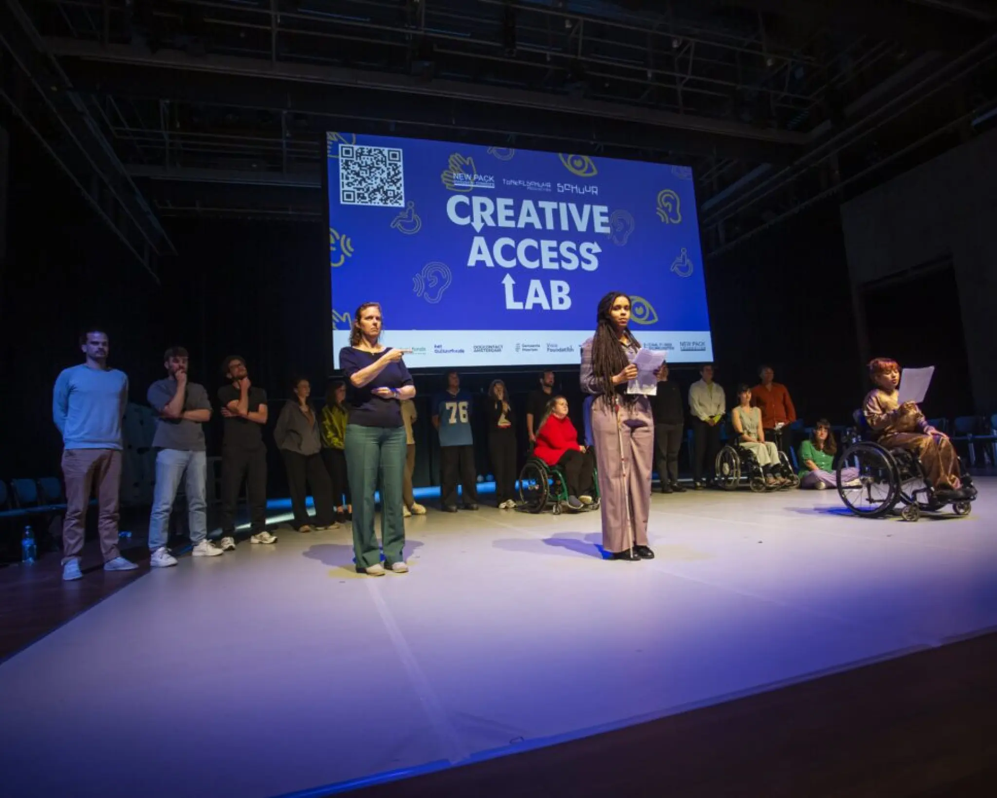 Presentatie van het manifest van Creative Access Lab. Een groep mensen staat op een rij aan de achterkant van het podium. Boven hen hangt een scherm waarop een presentatie te zien is met de tekst 'Creative Access Lab' en een QR-code. Op de voorgrond staan drie vrouwen. De linkervrouw is een tolk Nederlandse Gebarentaal. De middelste vrouw heeft een tekstin haar handen en heeft een taststok bij zich. De rechtervrouw zit in een rolstoel en heeft ook een tekst in haar hand.