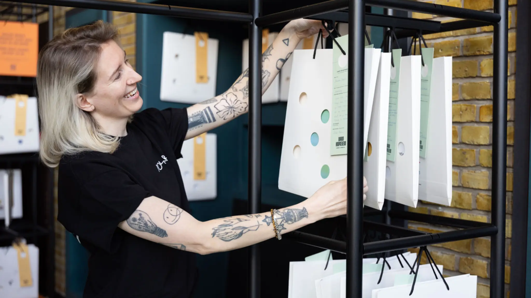 Een medewerker van de bibliotheek hangt lachend een wit tasje met een verhaal aan het rek. Ze draagt een zwart t-shirt en heeft verschillende tattoeages op haar armen.