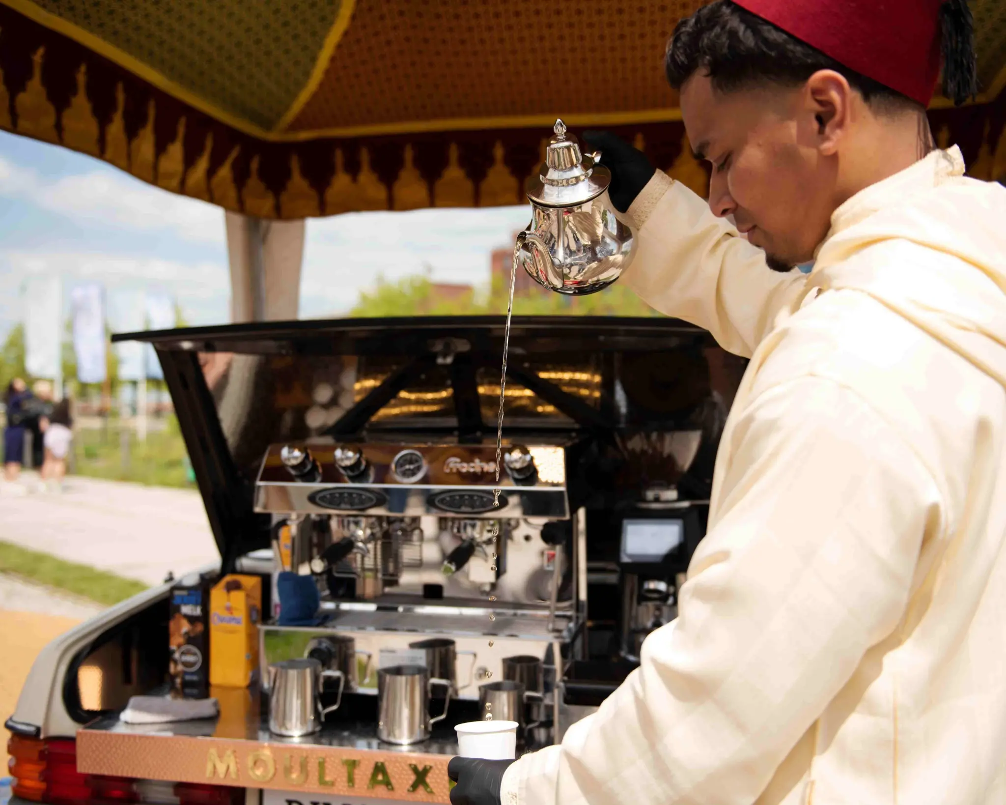Een man schenkt op traditionele wijze thee in. Hij draagt een rode fez en een witte djellaba. Naast hem is een koffiemachine te zien.