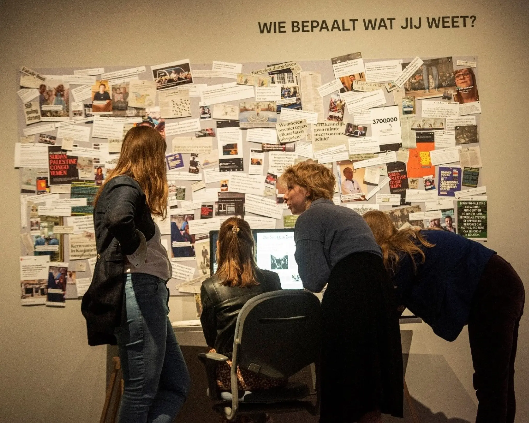 Vier jonge vrouwen staan en zitten bij een muur met krantenknipsels. Er staat een bureau met een pc voor. Boven de krantenknipsels staat Wie bepaalt wat jij weet?