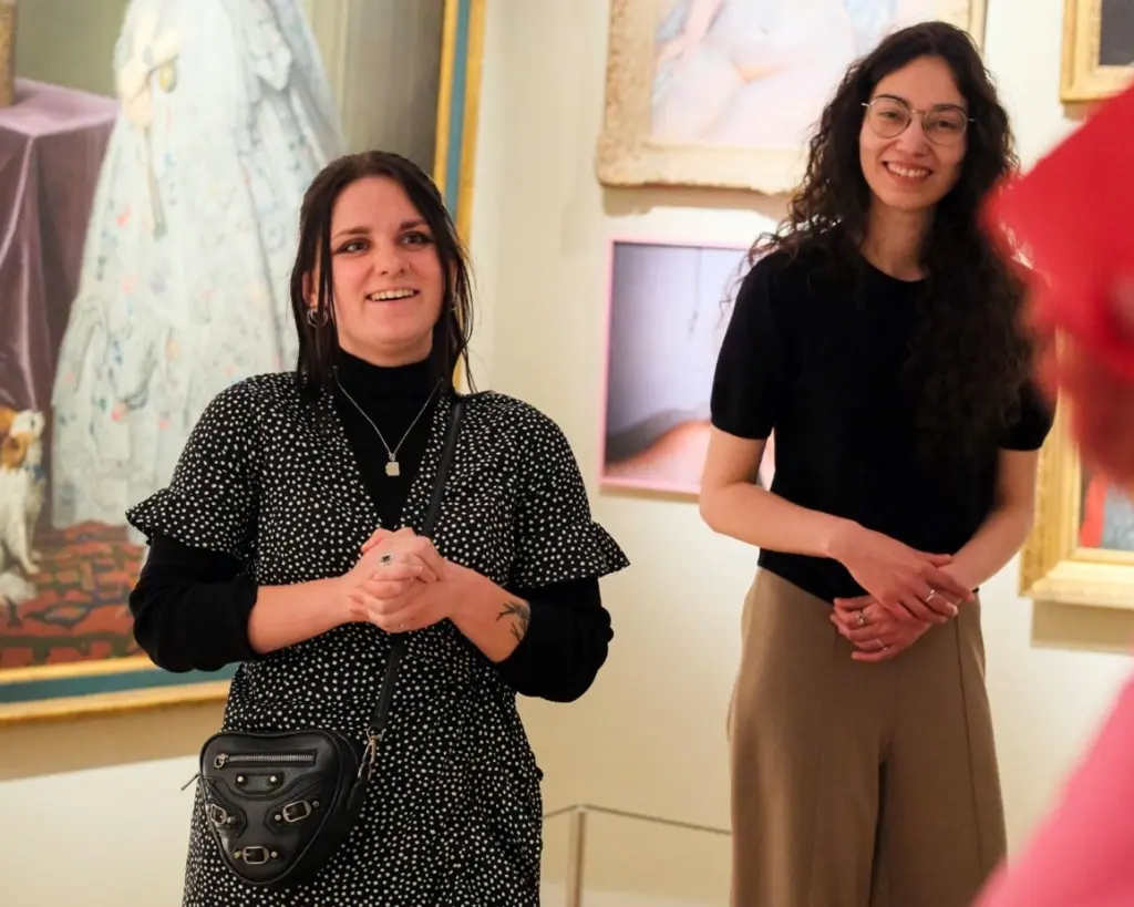 Twee vrouwen in een museumzaal. Ze kijken naar de camera. Achter hen zijn delen van schilderijen te zien Op de voorgrond is nog net een hand met een rood kaartje in beeld.