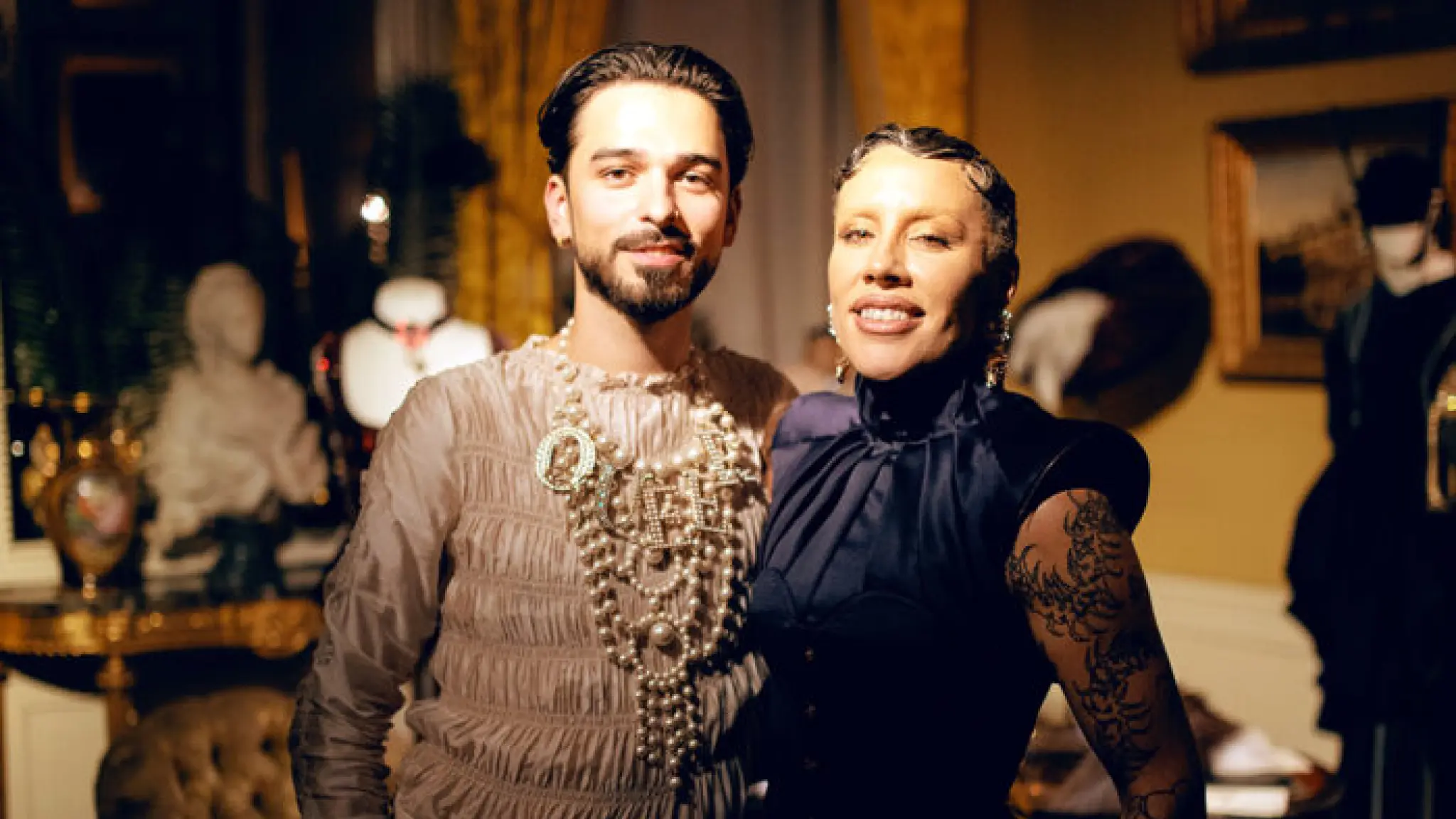 Twee mensen poseren glimlachend in een elegant ingerichte historische kamer. Ze dragen stijlvolle, opvallende kleding en staan voor een achtergrond met antieke meubels, kunstwerken en warme verlichting.