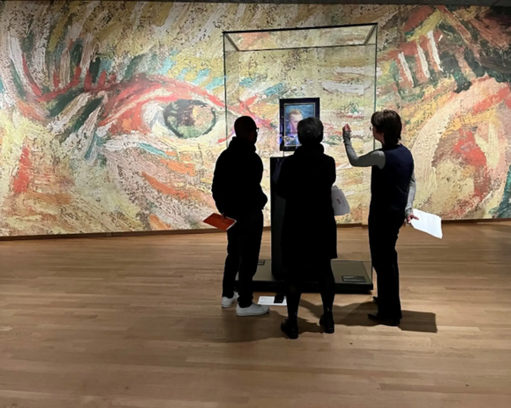 Drie mensen staan voor een vitrine met daarin een schilderij van Van Gogh. Op de achtergrond is een wand te zien met daarop een deel van een van de schilderijen van Van Gogh.  Je ziet een oog en een oor.