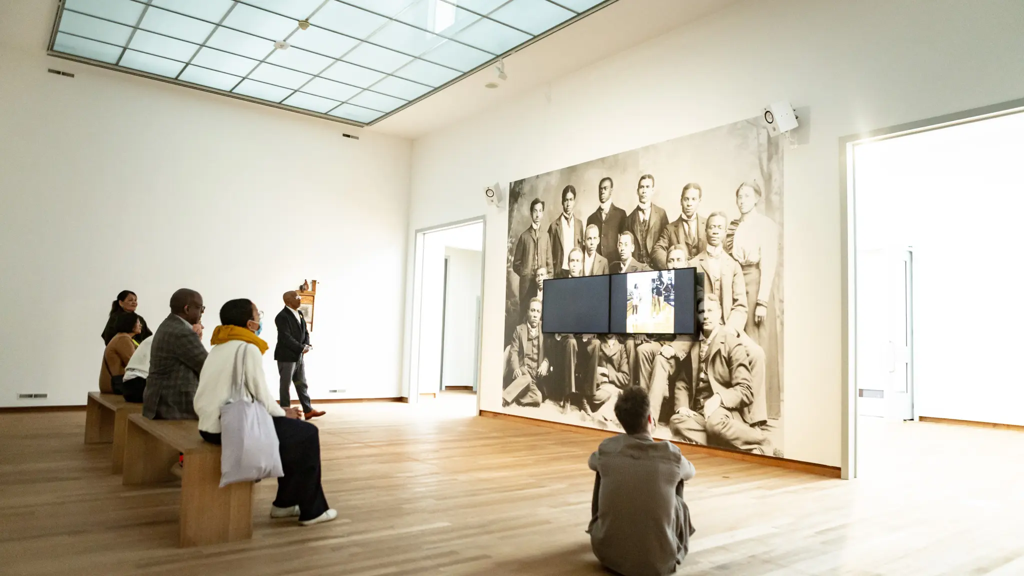 Bezoekers zitten en staan in een lichte museumzaal en bekijken een videopresentatie. Op de muur achter de schermen staat een grote zwart-witfoto van een groep mensen. De ruimte heeft een houten vloer, hoge plafonds en veel daglicht.