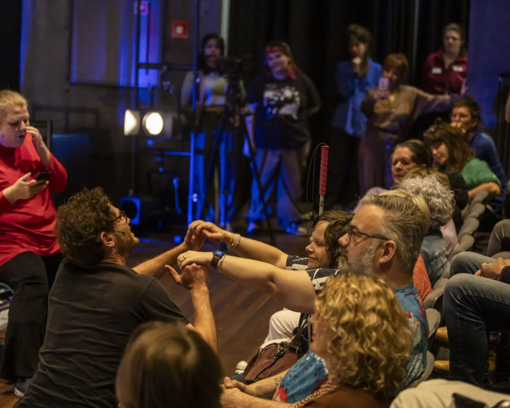 Impressie van de slotavond van Creative Acceess Lab. Op het podium zit een vrouw in een rolstoel. Zij leest iets voor van haar mobiel. Een amn op het podium pakt de handen vast van iemand in het publiek. Op de achtergrond maken mensen foto's met de camera van hun telefoon.