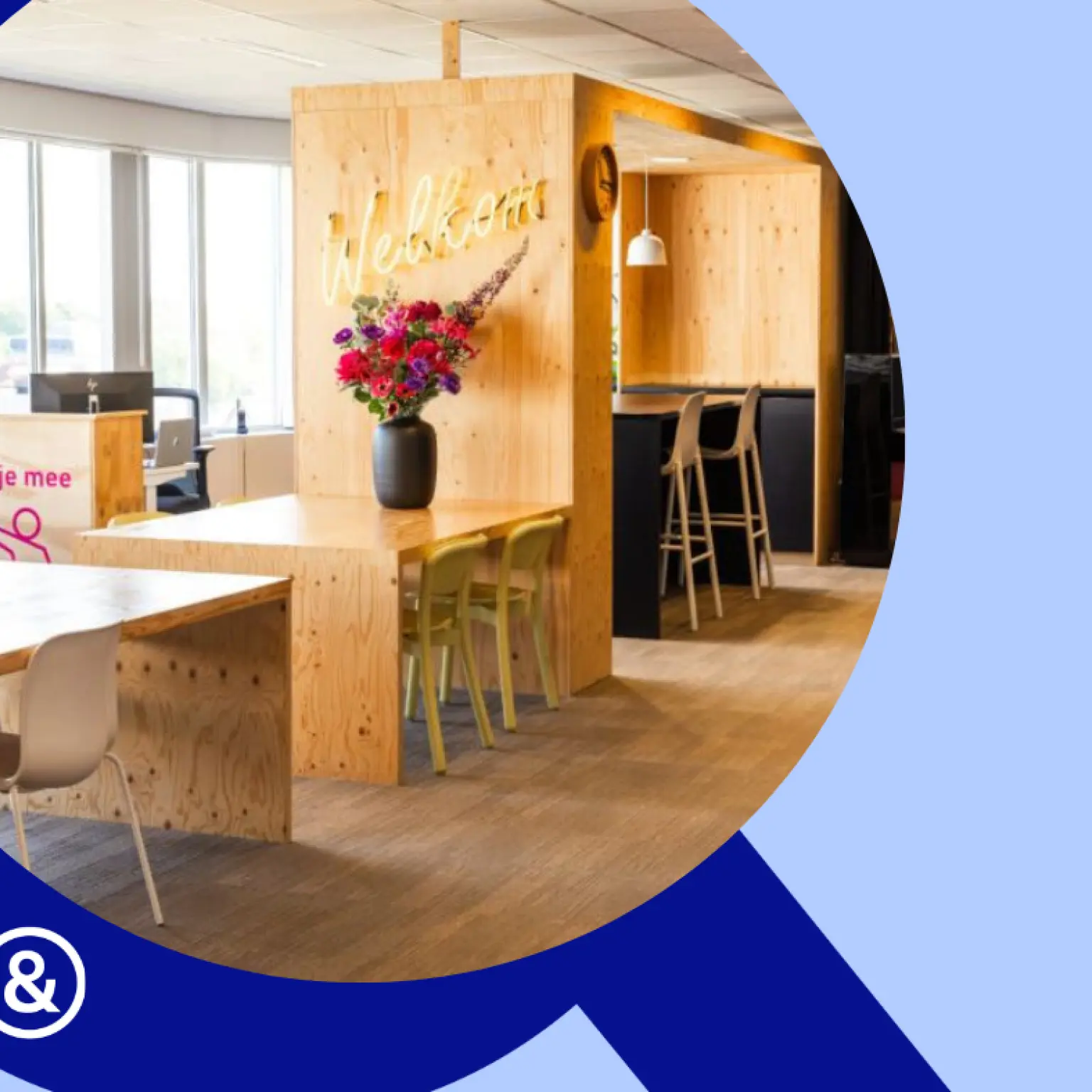 Foto van een open werkruimte met houten tafels en bureaus. Op de voorkant van de receptie staat de tekst "Waar kan ik je mee helpen?". Op een tafel staat een bos bloemen met aan de wand erboven de tekst "Welkom"