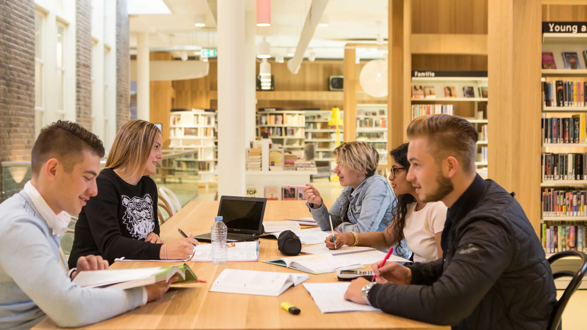 Vijf jonge mensen zitten aan tafel in een bibliotheek. Op tafel liggen schrijfblokken, boeken, een laptop en schrijfwaren. Om hen heen zijn kasten vol boeken te zien.