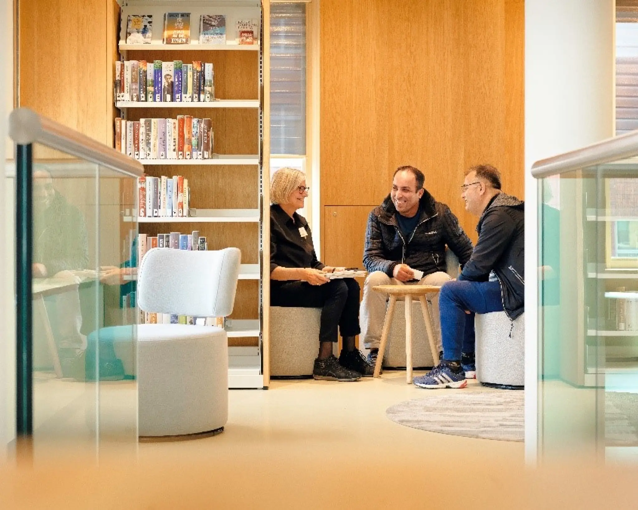 Drie mensen zitten lachend op krukjes in een hoekje van de bibliotheek. Tussen hen in staat een krukje. Naast hen is een kast met boeken te zien.
