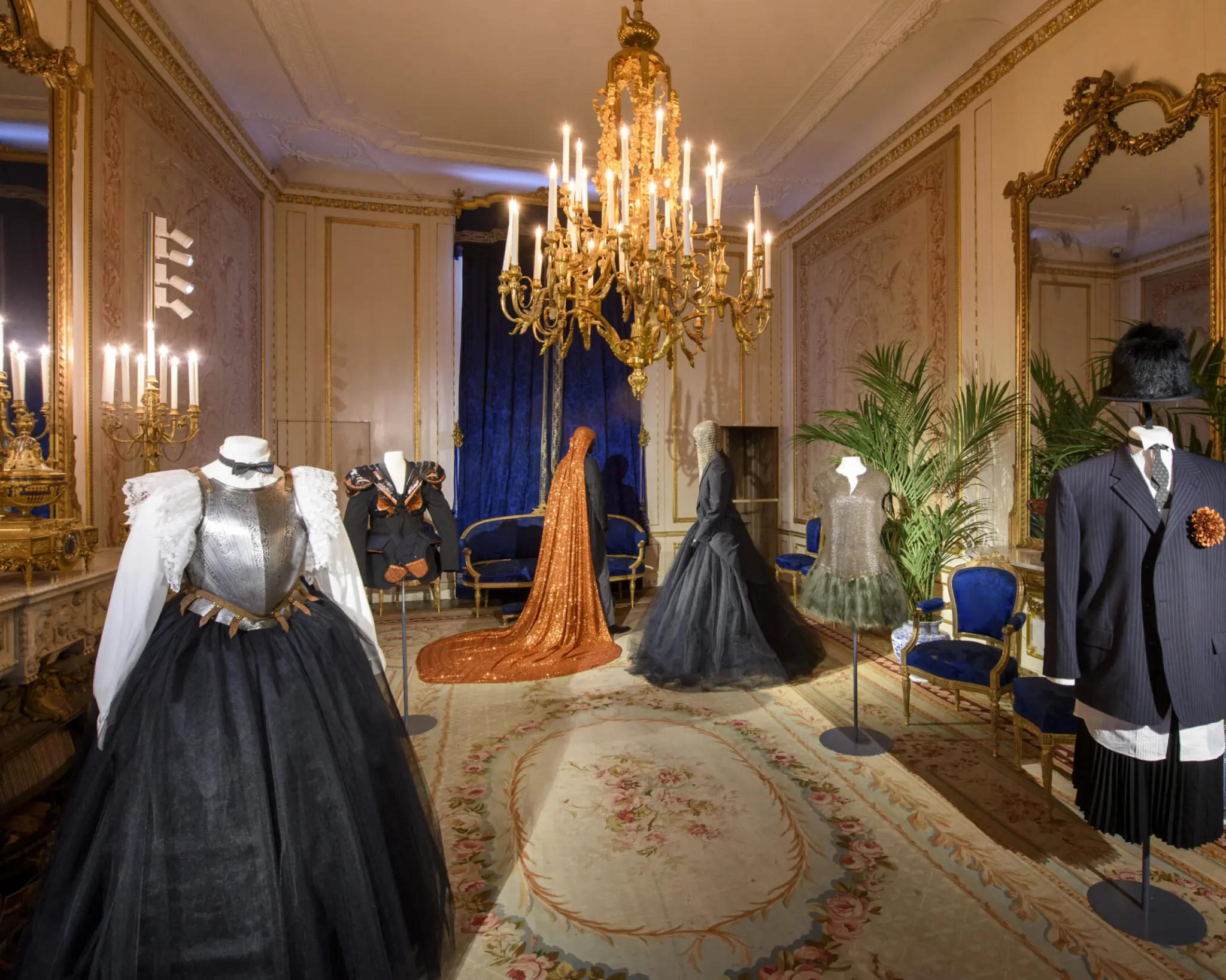 Historische salons met kroonluchters en spiegels, ingericht als mode-expositie met opvallende kledingstukken op paspoppen.